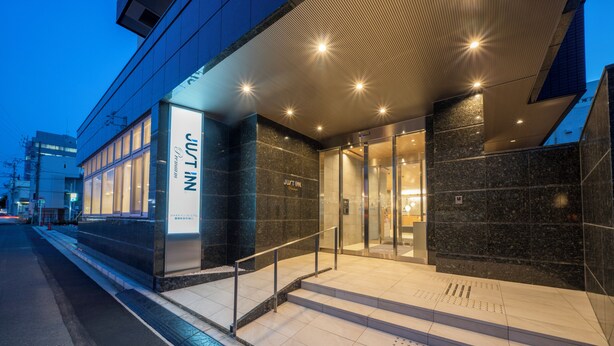 Una entrada de hotel moderna con fachada de vidrio, una escalera visible y un letrero que dice 'JUST IN'.