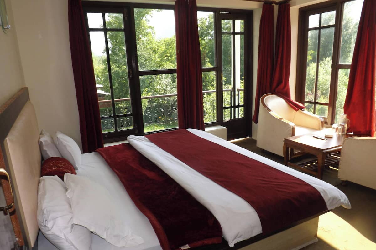 A hotel room with a bed, a desk with a chair, a window with curtains, and a view of greenery outside.