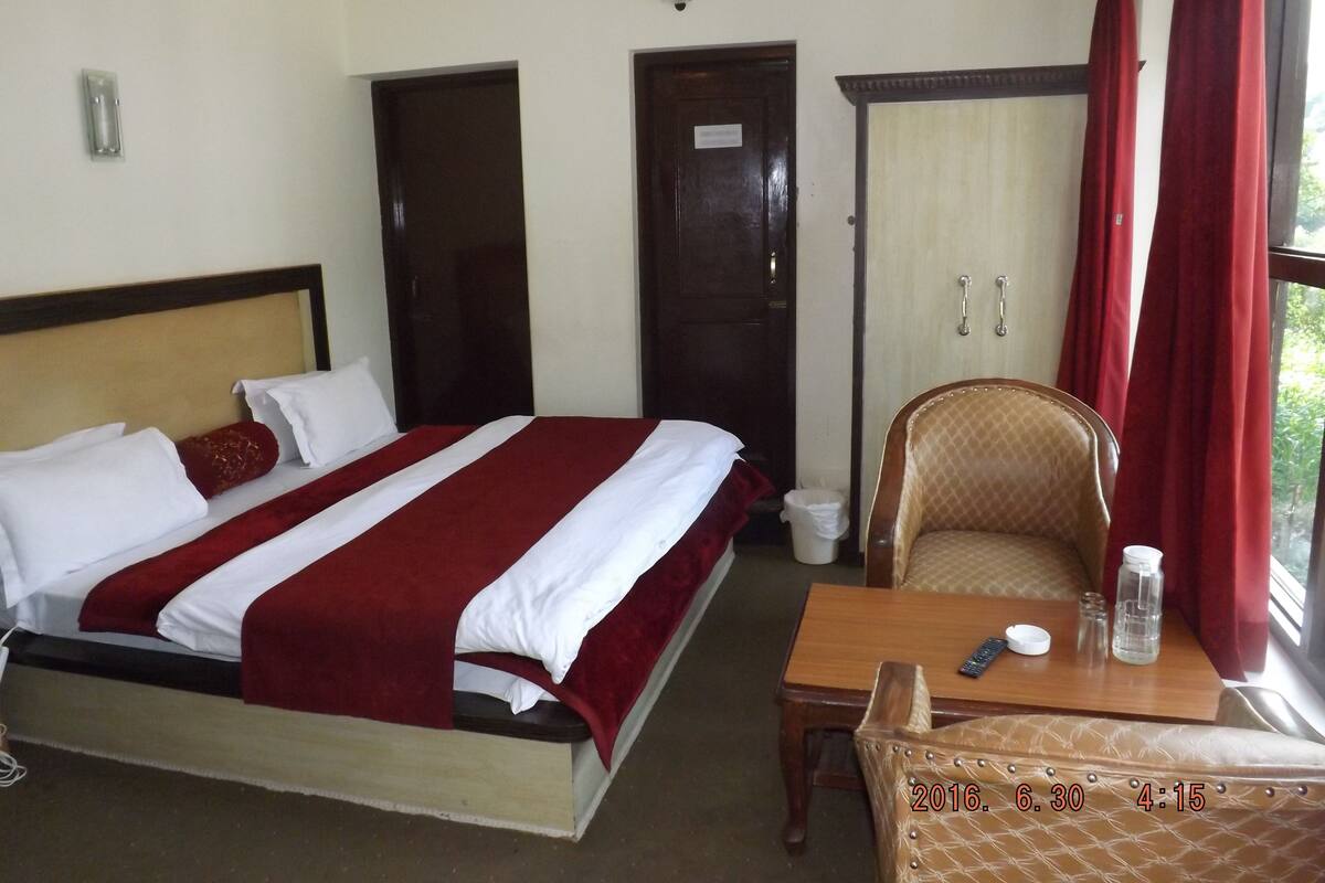 A hotel room with a bed, a wooden chair, a small table, and a window with red curtains.
