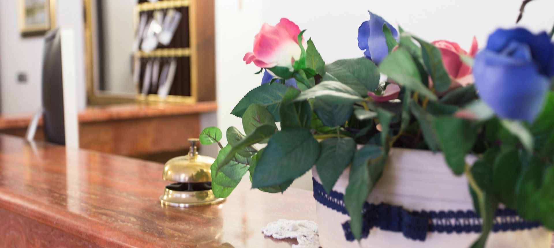 Una mesa de madera con una campana de latón, una planta en maceta con rosas y un florero blanco con un diseño azul.