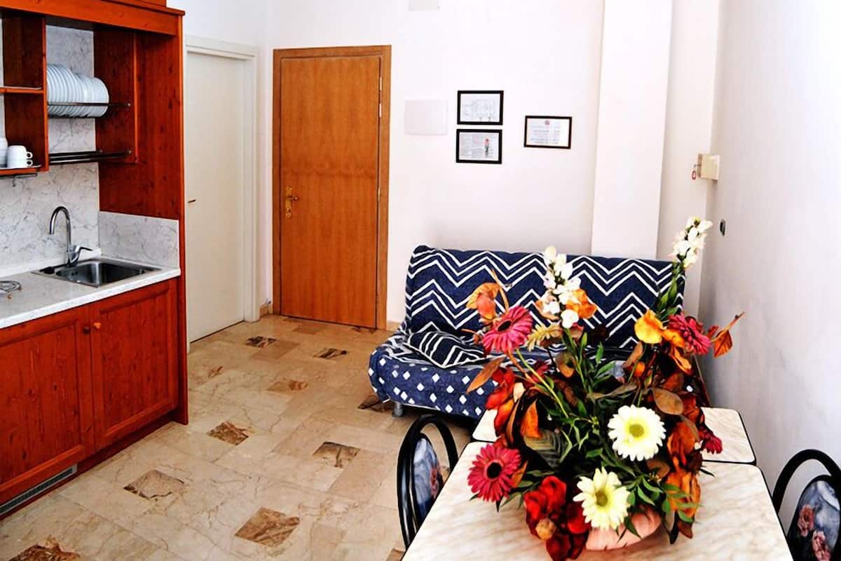 A compact kitchen with a wooden cabinet, a sink, and a door. A patterned sofa and a table with a floral arrangement are in the living area.