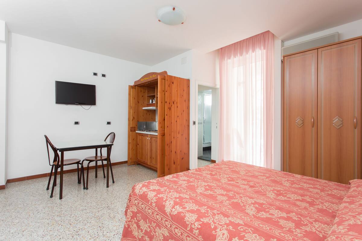 A bedroom with a bed, a wooden wardrobe, a television, a desk with chairs, and a bathroom door.