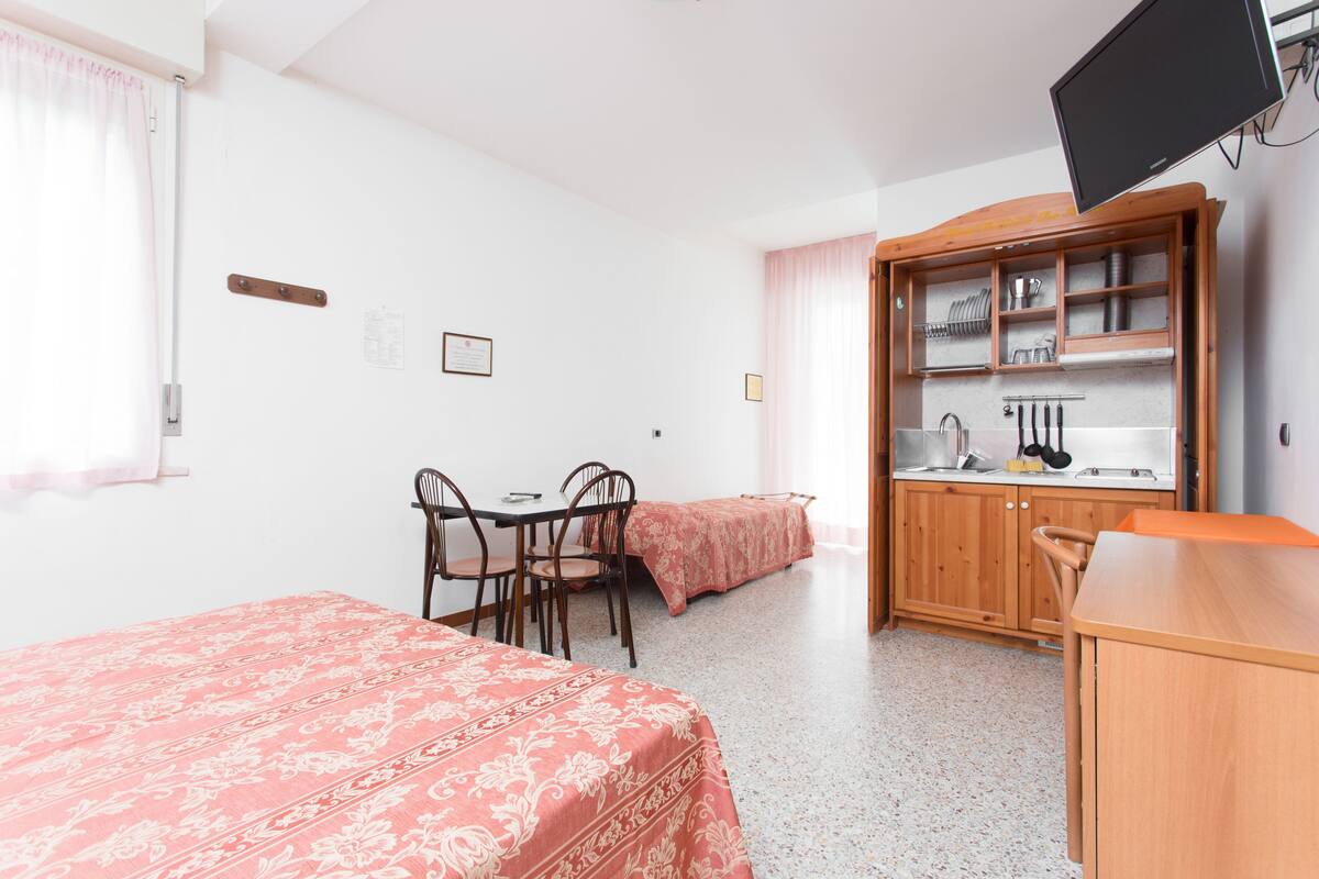 A room with two beds, a dining table with chairs, a kitchen area with cabinets, and a television mounted on the wall.