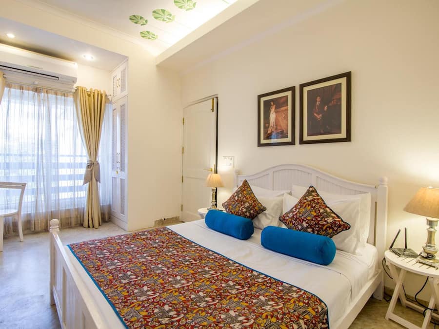 Quarto de hotel com cama, uma cadeira, uma mesa com abajur e duas fotos emolduradas na parede.