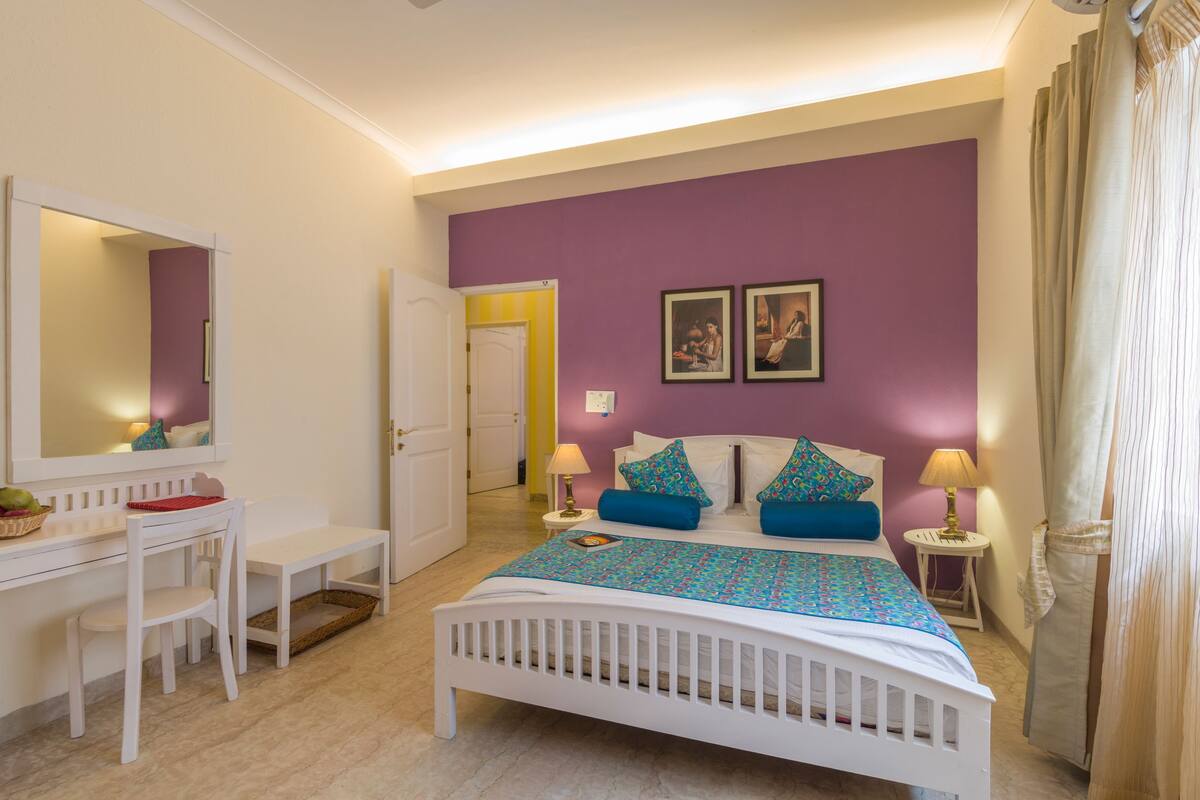 A bedroom with a white bed, purple walls, a desk with a chair, and a small table with a lamp.