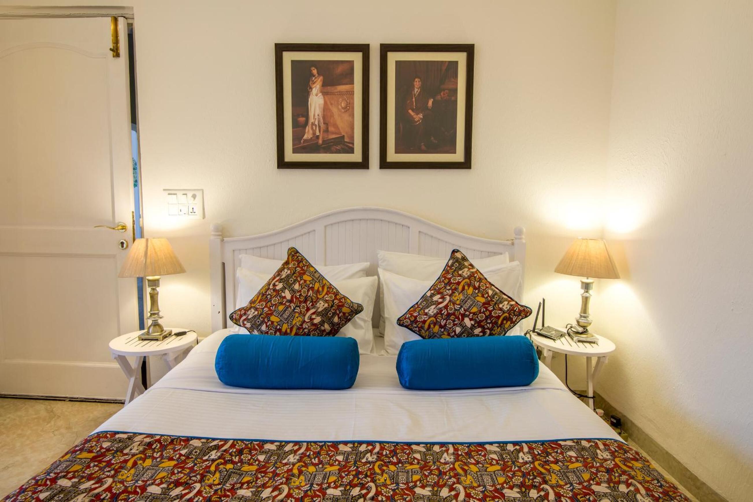 A neatly made bed with two blue pillows and a patterned bedspread, flanked by two bedside tables with lamps, in a room with a white headboard and two framed pictures on the wall.