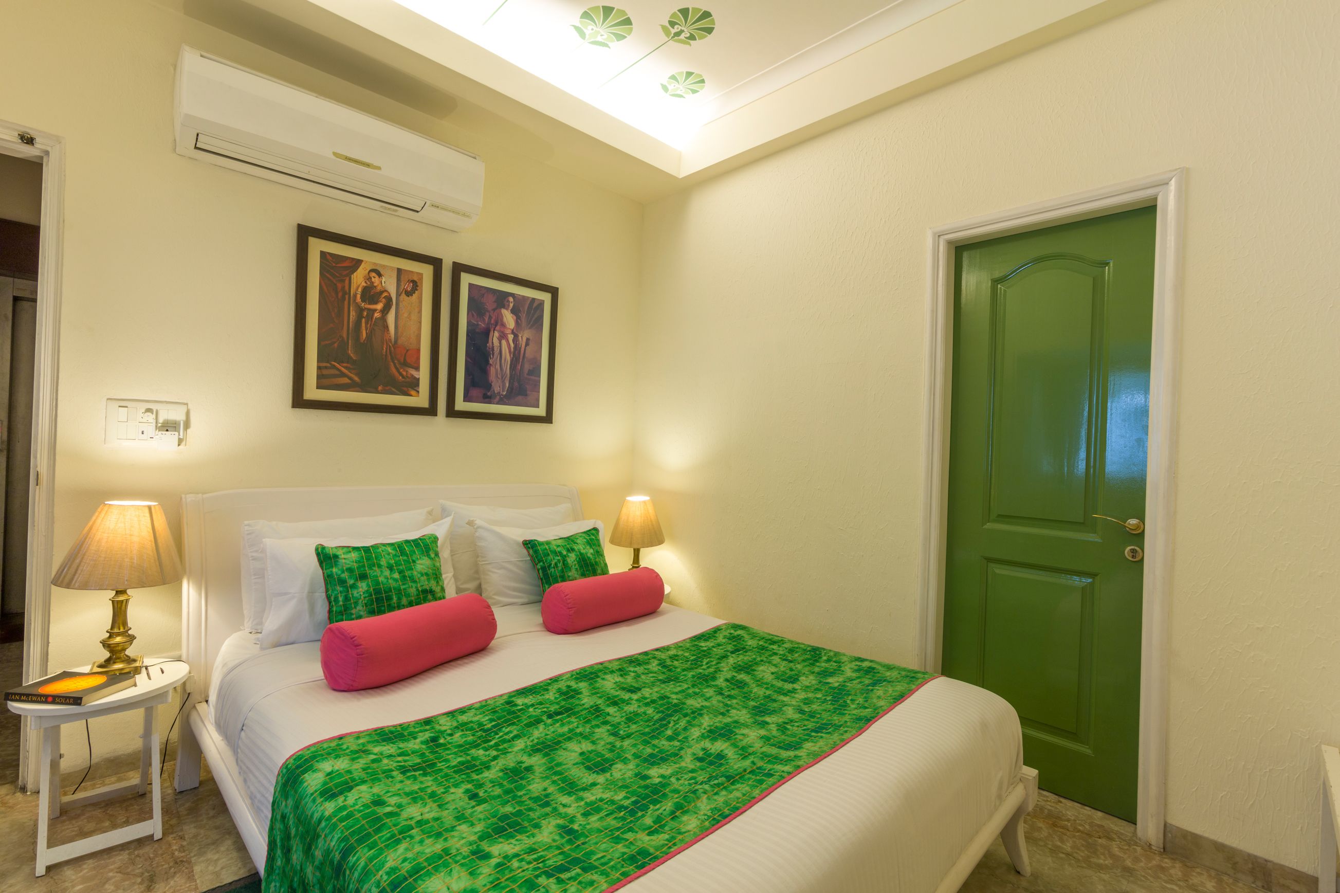 A hotel room with a bed, two wall-mounted paintings, a green door, and an air conditioning unit.