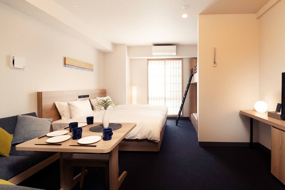 Quarto de hotel com cama, escrivaninha com televisão de tela plana, sofá e pequena mesa com xícaras e um vaso.