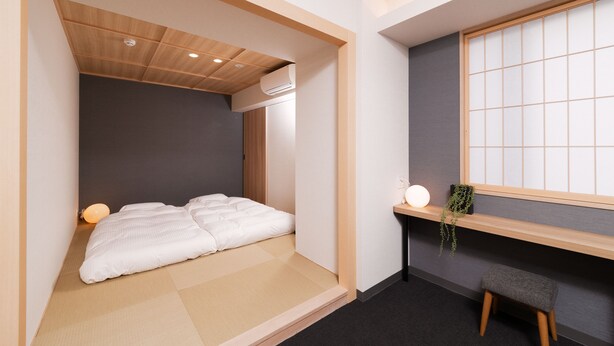 Una habitación pequeña con cama, un taburete, un estante con una planta y una ventana con diseño de cuadrícula.