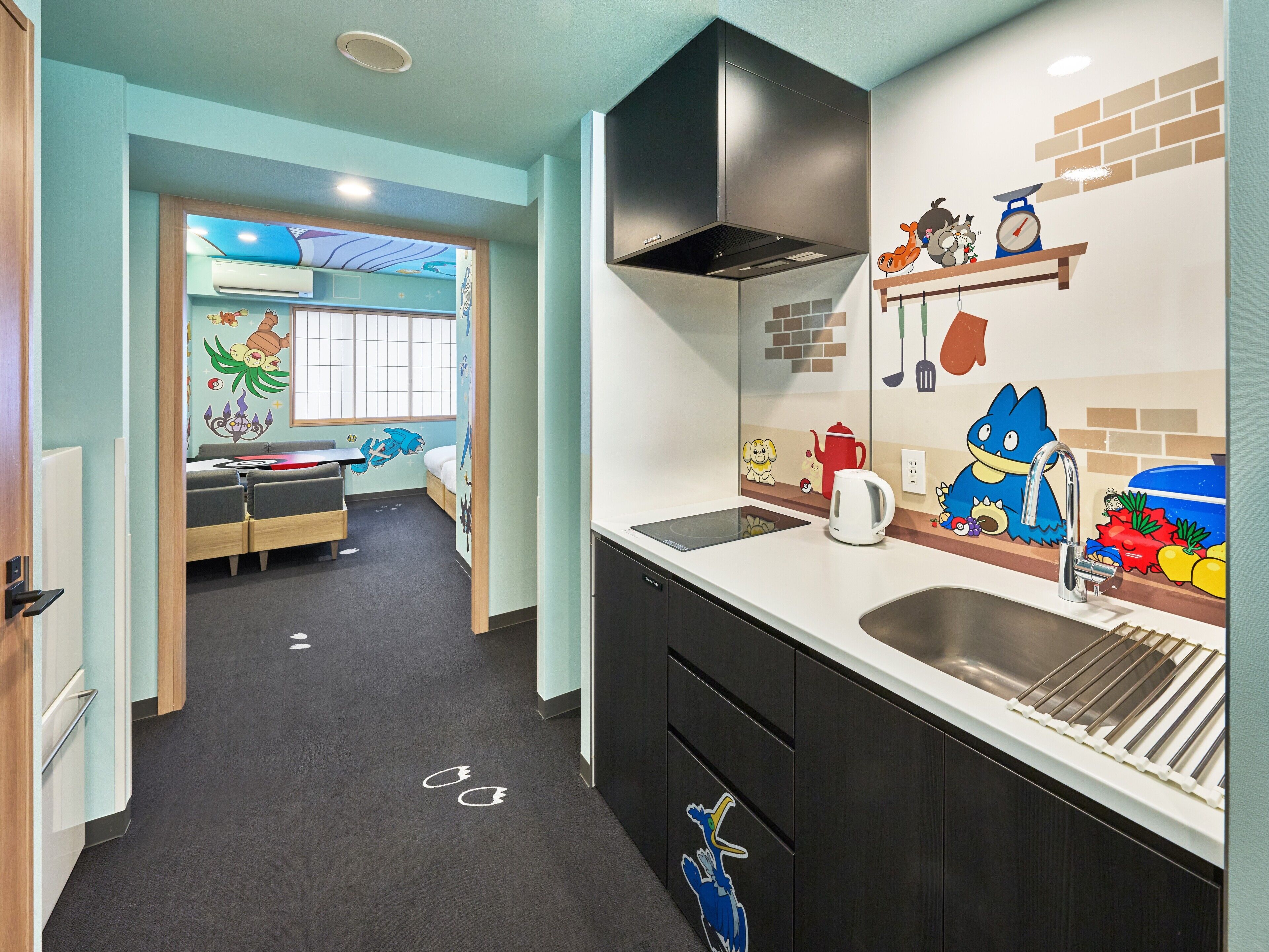 A modern kitchen with a black range hood, a white kettle, and a sink with a faucet. There are cartoon characters on the wall, and a window with a view of a room with a bed and a desk.