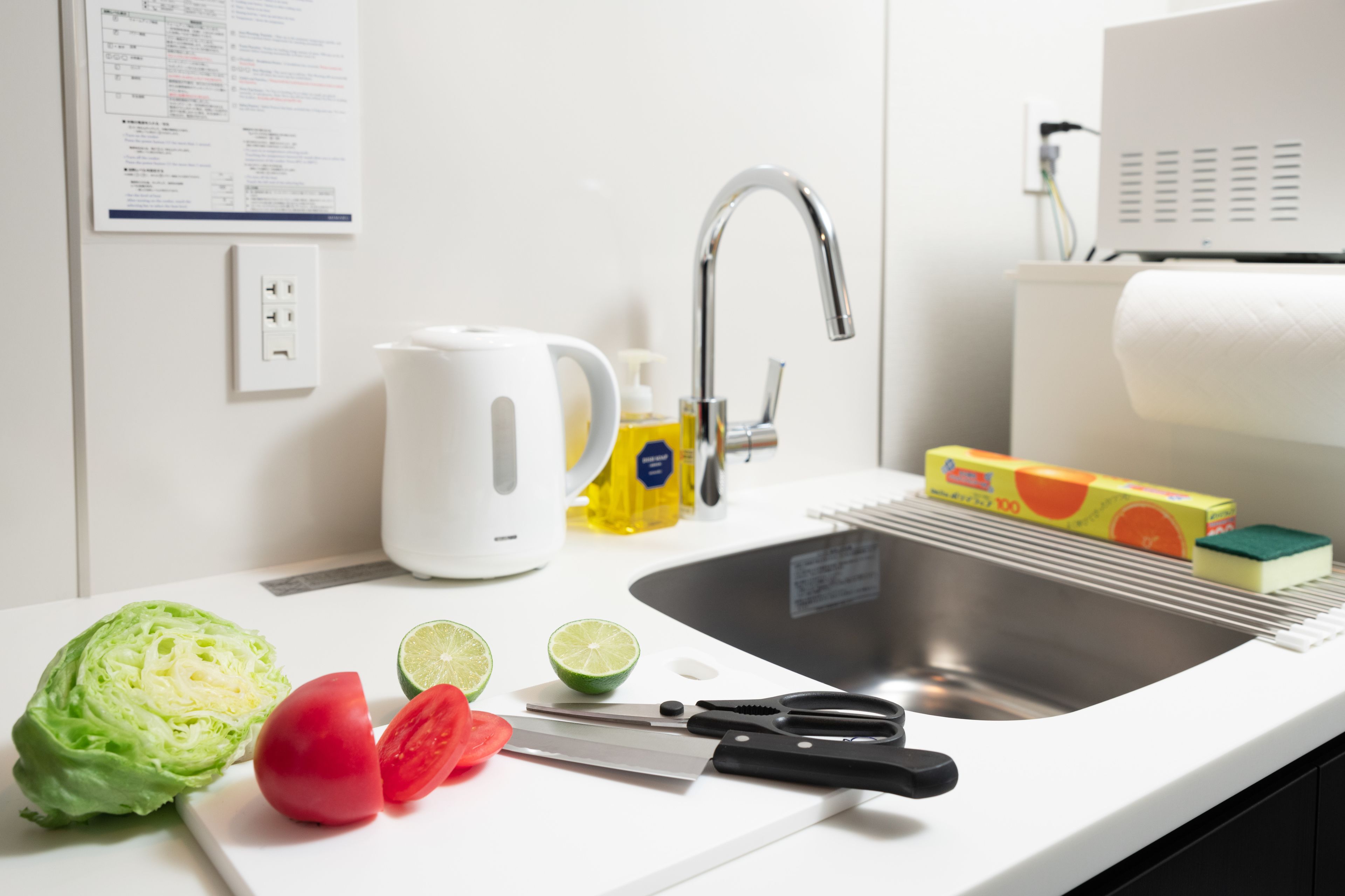 A kitchen with a sink, a cutting board with vegetables, a kettle, and a soap dispenser.