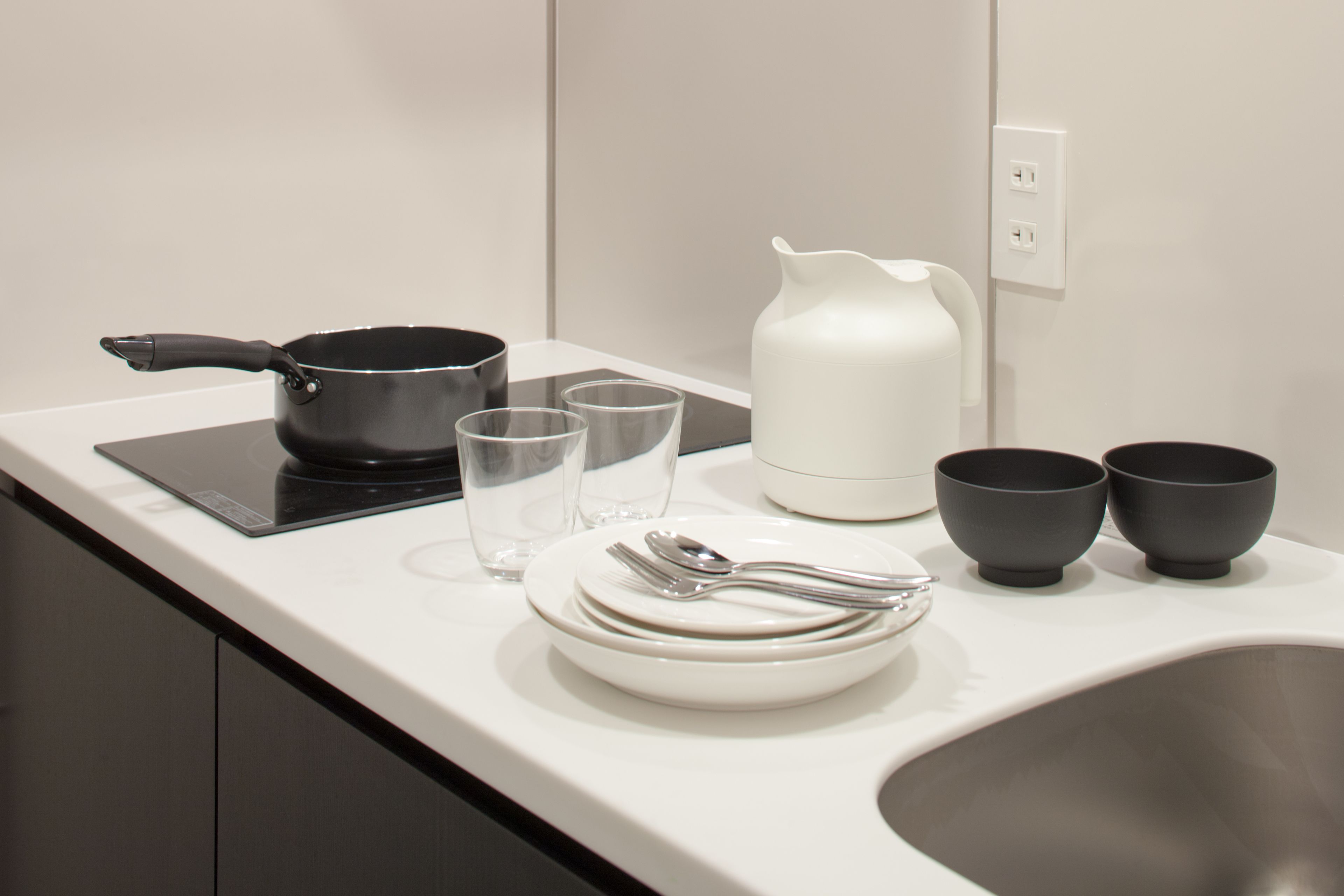 A kitchen counter with a stove, a kettle, two cups, a plate with cutlery, and a sink.
