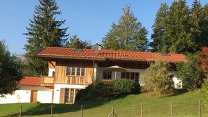 A two-story wooden house with a red-tiled roof, surrounded by a grassy area and trees. - Bergblick Riedlern, Kreuth Riedlern - Mountain View Riedlern - Apartment (Kreuth)