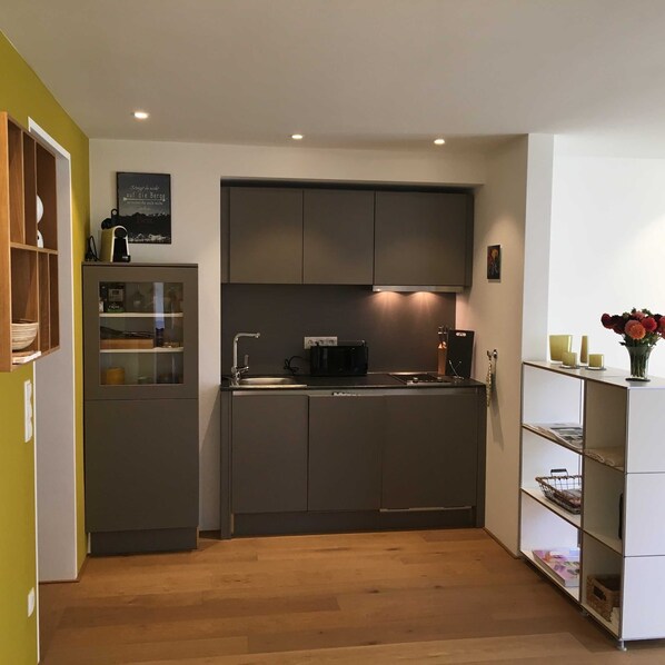 A compact kitchen with a built-in oven, a sink, and a countertop. There is a small cabinet with a glass door and a shelf with decorative items. - Bergblick Riedlern, Kreuth Riedlern - Mountain View Riedlern - Apartment (Kreuth)