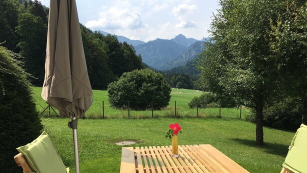 A wooden table with a vase of flowers, two chairs, and an umbrella in a grassy outdoor area with a mountain view. - Bergblick Riedlern, Kreuth Riedlern - Mountain View Riedlern - Apartment (Kreuth)
