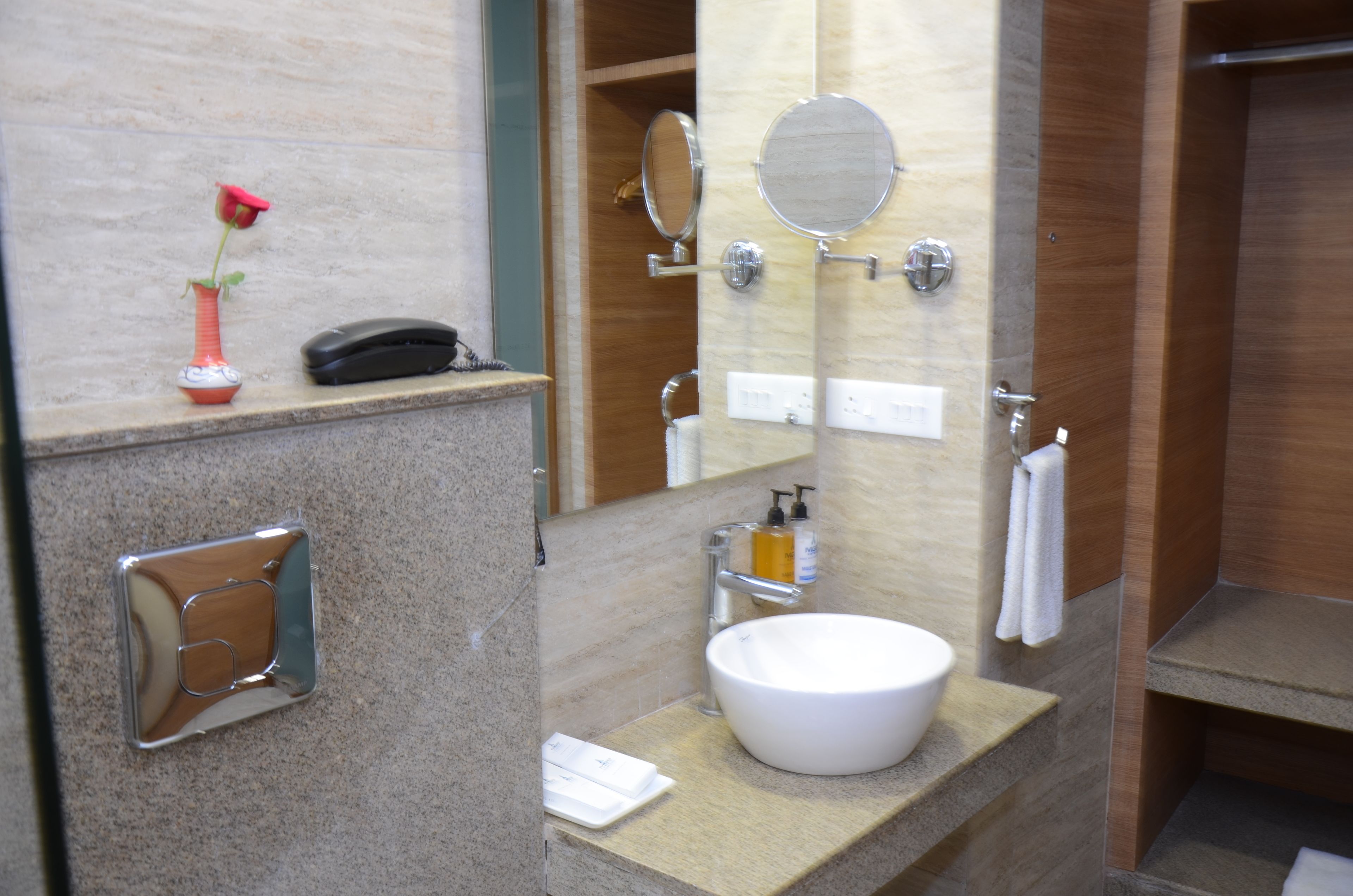 A bathroom with a white sink, a mirror, a telephone, and a rose vase.
