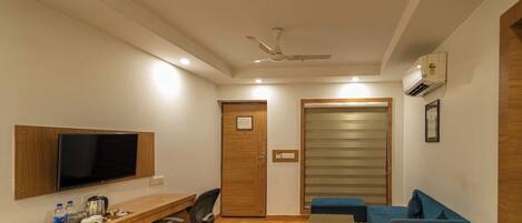 A hotel room with a wooden desk, a flat-screen TV, a blue sofa, a small table, and a ceiling fan.