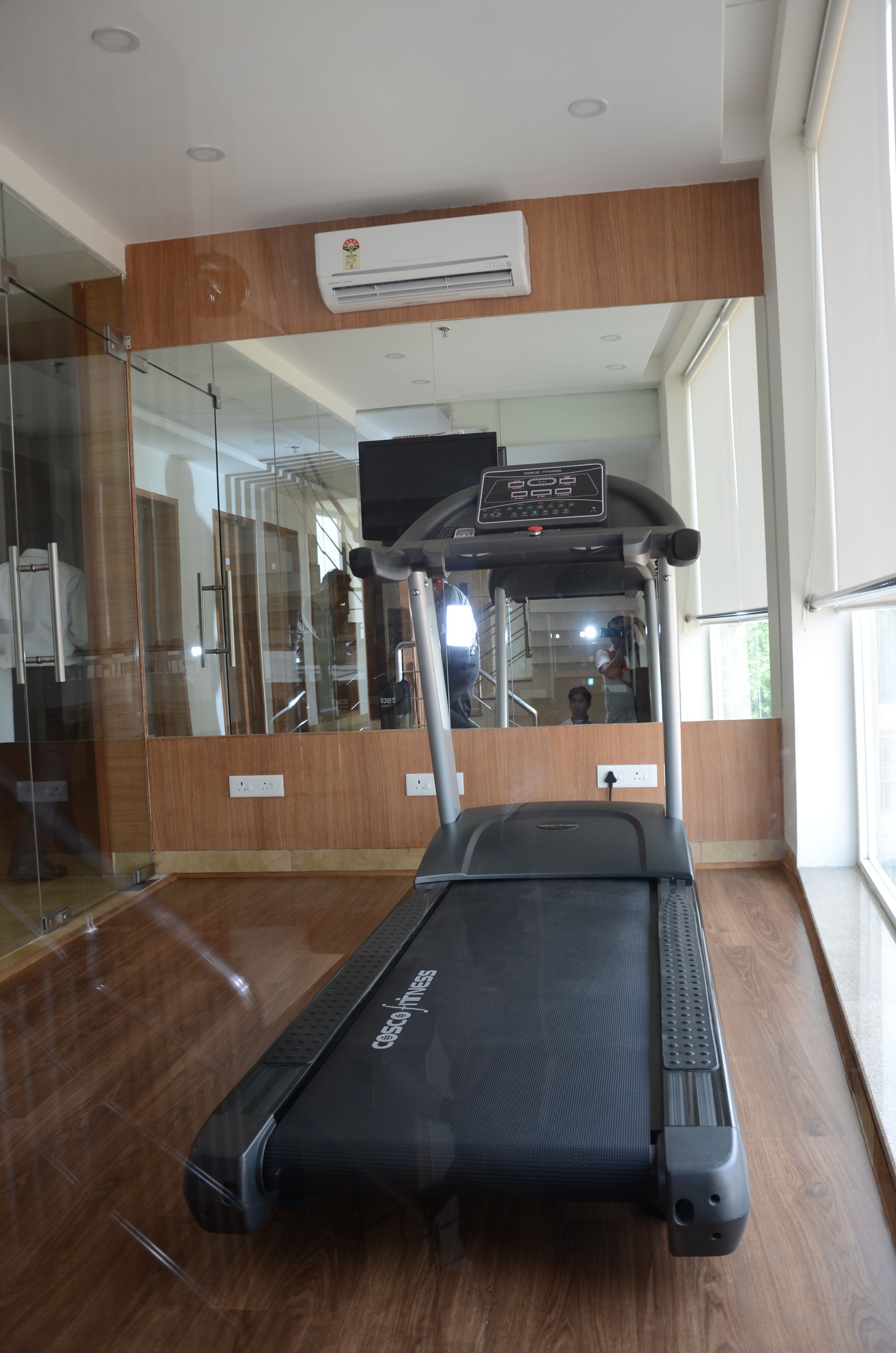 A treadmill in a room with a wooden floor, a glass wall, and a large mirror.