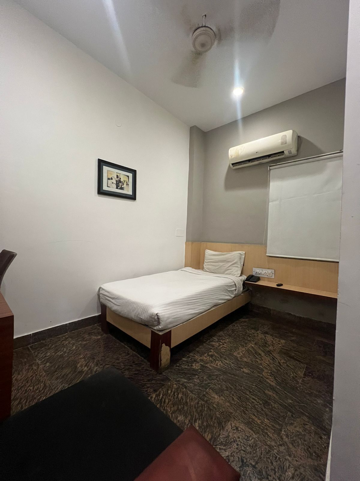 Una habitación de hotel pequeña y ordenada con una cama individual, una mesita de noche, una silla, un equipo de aire acondicionado y un cuadro en la pared.