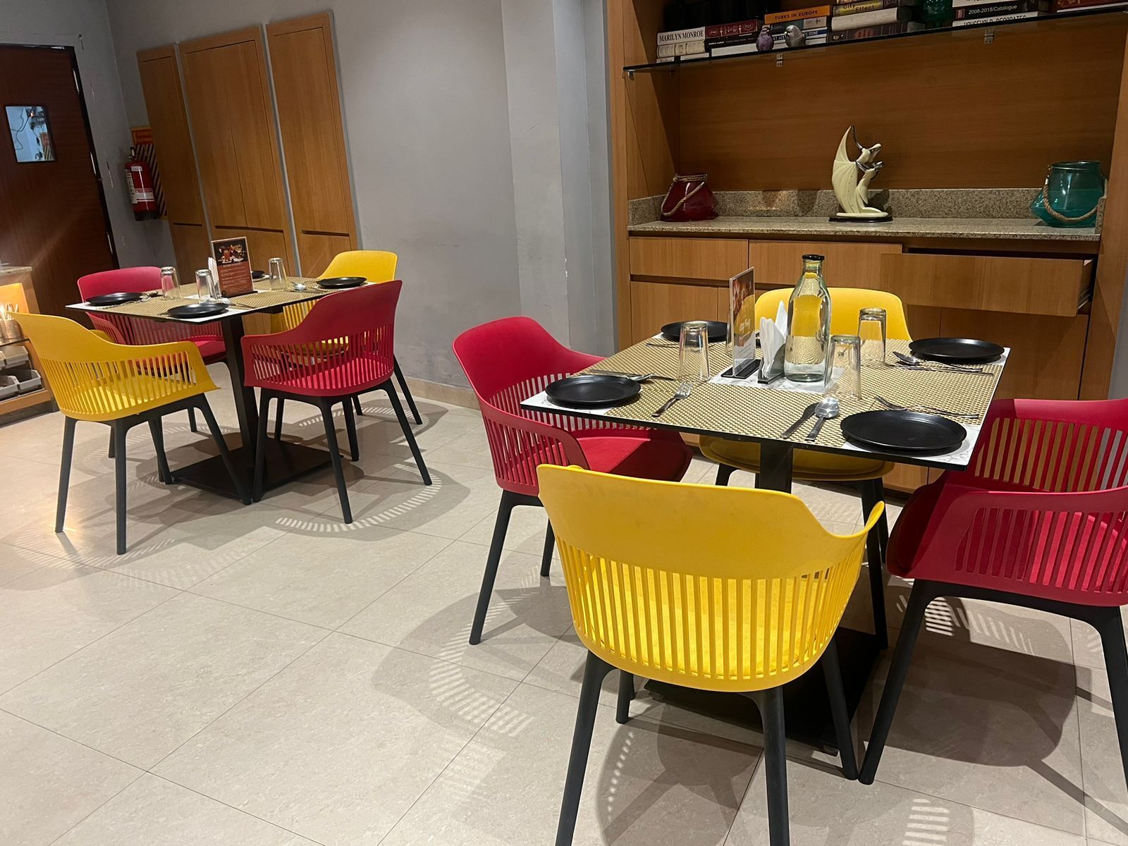 Un comedor con una mesa dispuesta para cuatro personas, rodeado de sillas coloridas, y una zona de cocina con armarios de madera y una estantería con libros y objetos decorativos.