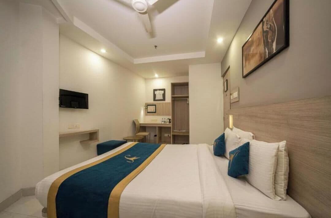 A hotel room with a bed, a desk, a chair, a television, and a bathroom visible in the background.