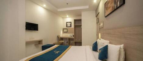 A hotel room with a bed, a desk, a chair, a television, and a bathroom visible in the background.