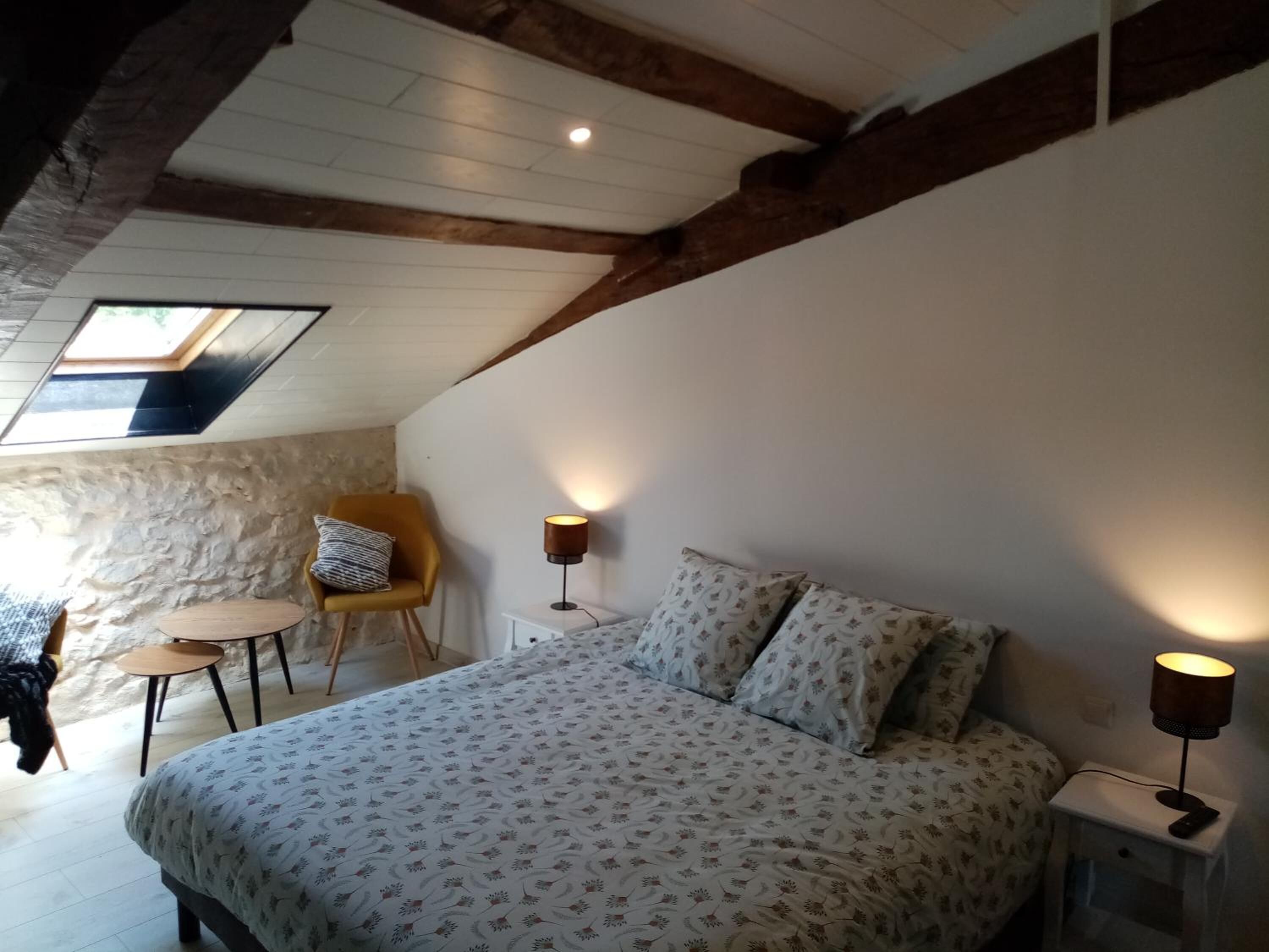 A bedroom with a bed, a chair, a small table, and a lamp. There is a skylight above the bed.