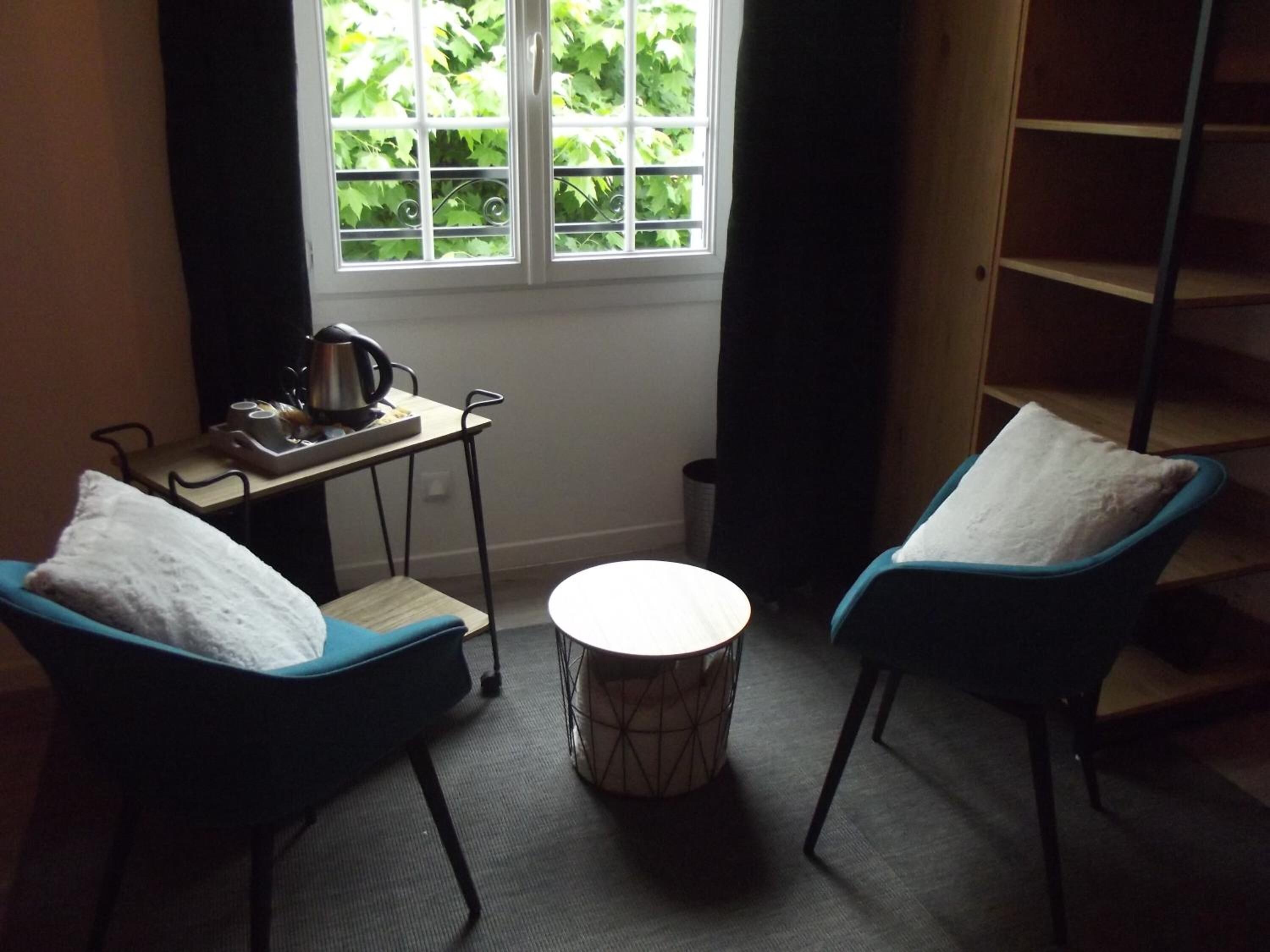 A room with two blue chairs, a small round table, a window with a view of greenery, and a metal shelf unit.