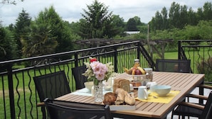 Un área de comedor al aire libre con una mesa de madera preparada para el desayuno, rodeada de sillas, y vista a un jardín y árboles al fondo.