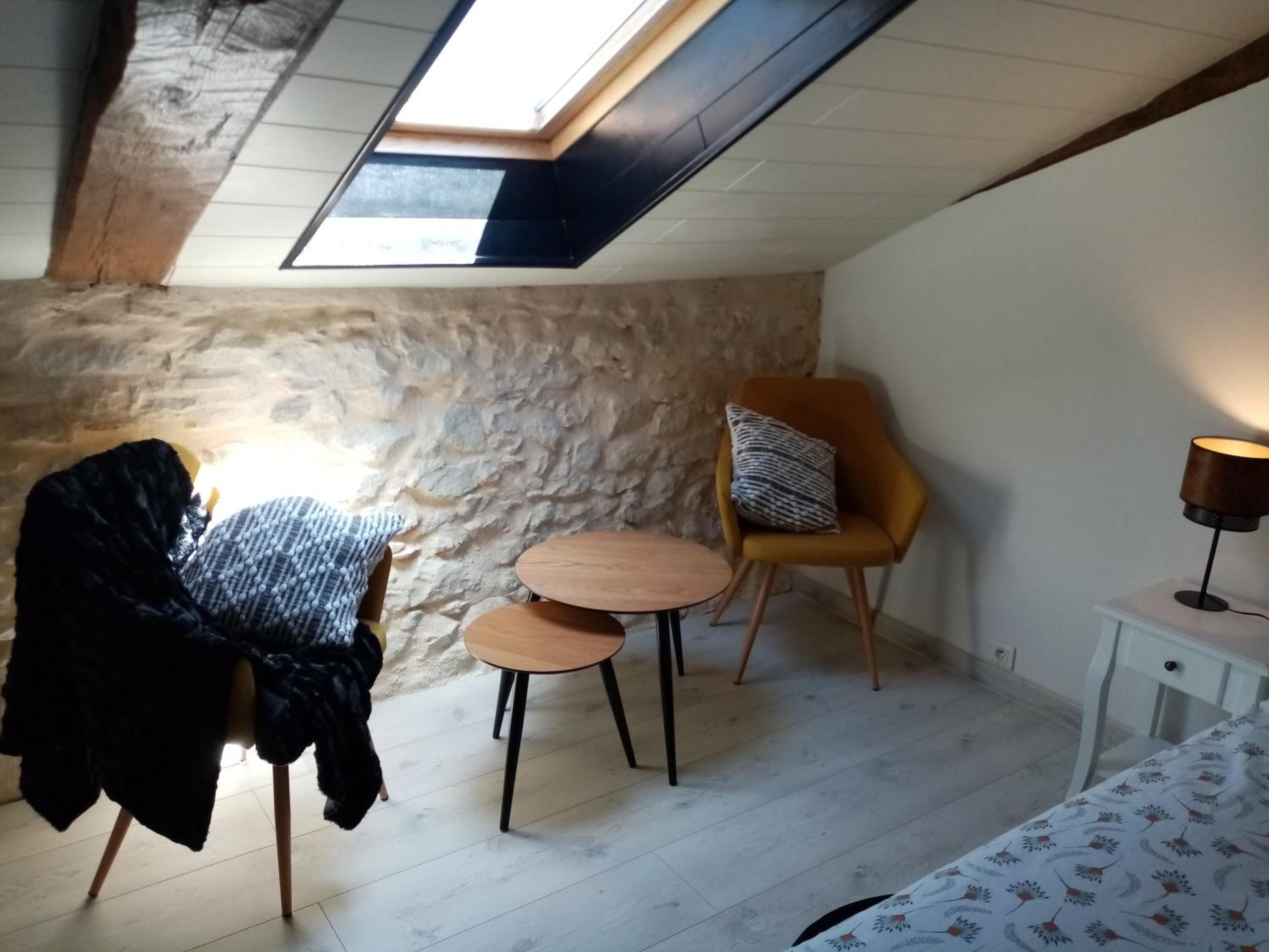 A cozy attic room with a stone wall, a wooden table with two chairs, a yellow armchair with a patterned pillow, a bedside table with a lamp, and a bed with a floral bedspread.