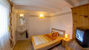 A bedroom with a bed, a bedside table with a lamp, a television, a mirror, and a bathroom with a sink. - Lindenschänke Hotel garni (Werl)