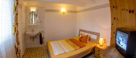 A bedroom with a bed, a bedside table with a lamp, a television, a mirror, and a bathroom with a sink.