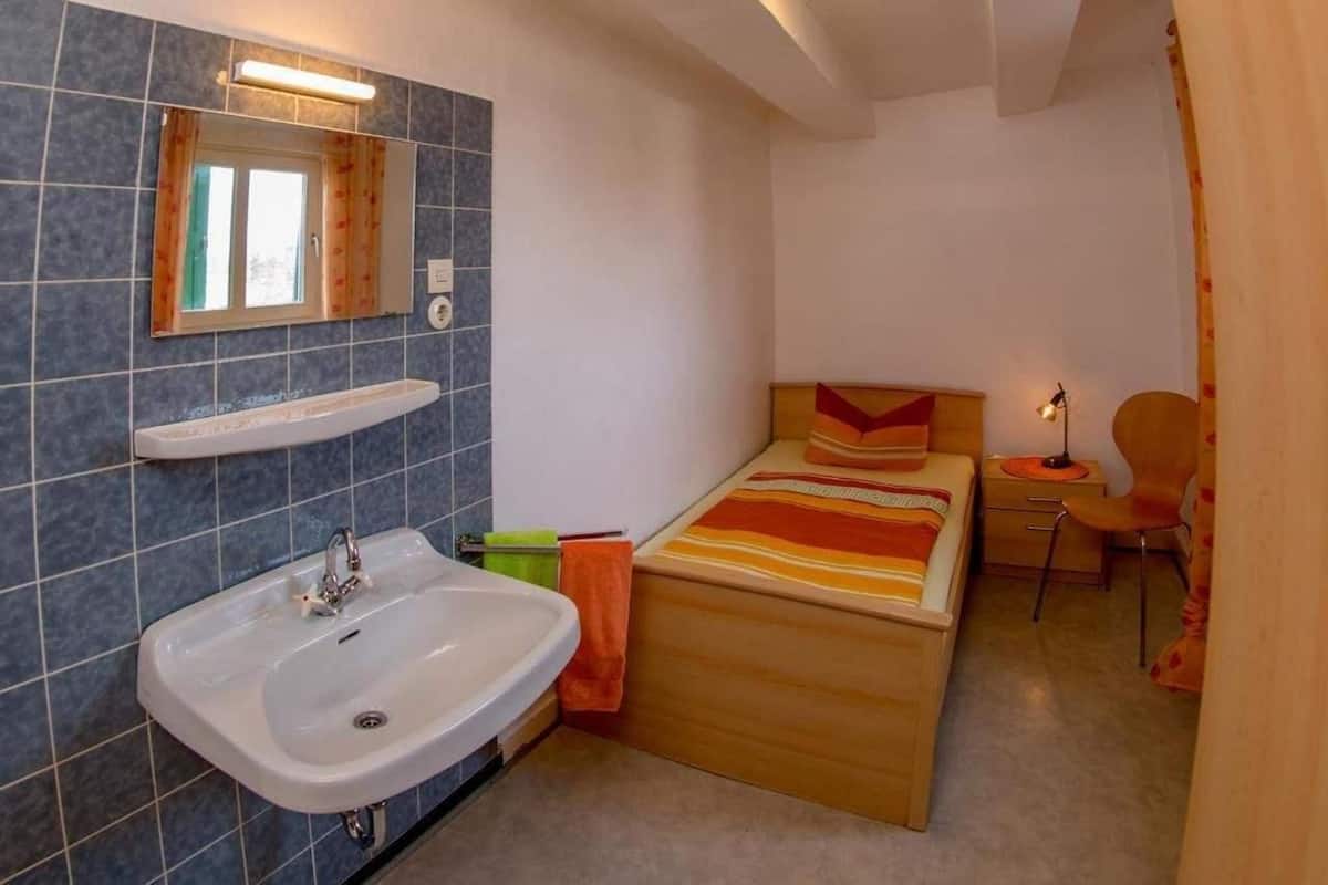 A small, compact bedroom with a single bed, a bedside table, a chair, and a small desk with a lamp. There is a bathroom with a sink and a mirror.