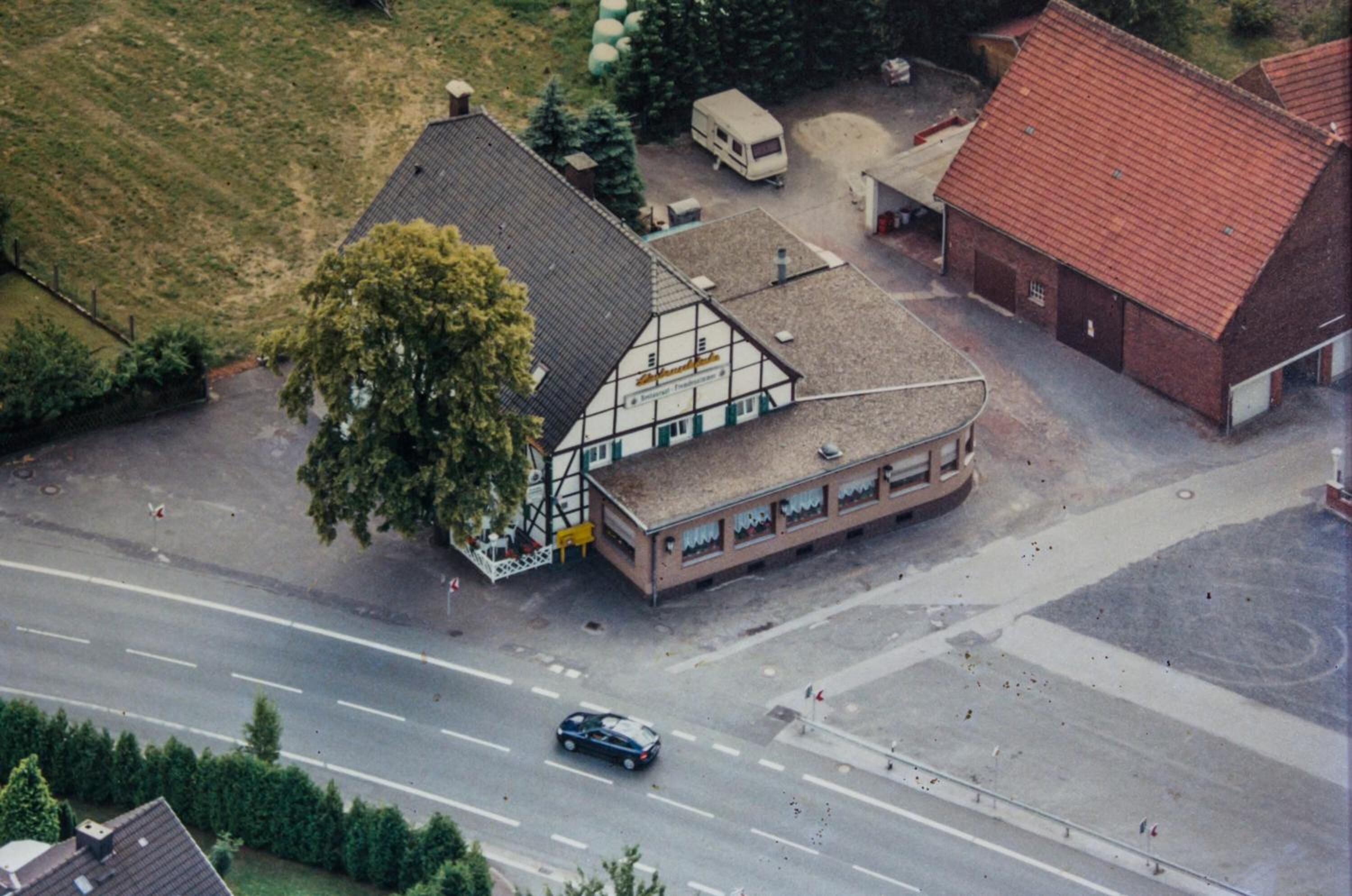 Vista aérea de un edificio con techo inclinado, un árbol grande al frente y una calle con un coche.