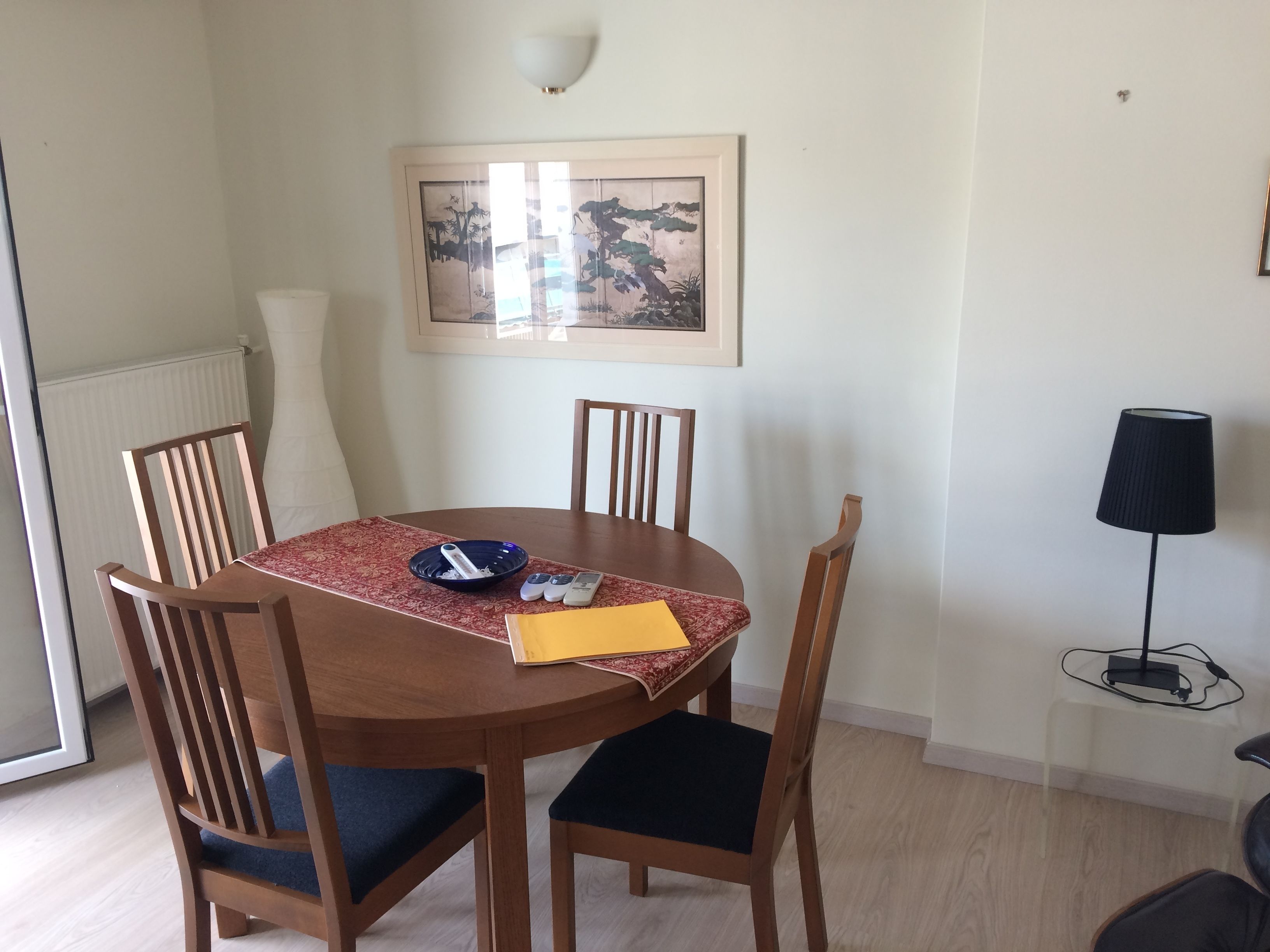 A dining area with a round wooden table, four chairs, a framed artwork on the wall, a radiator, and a lamp on a small table.