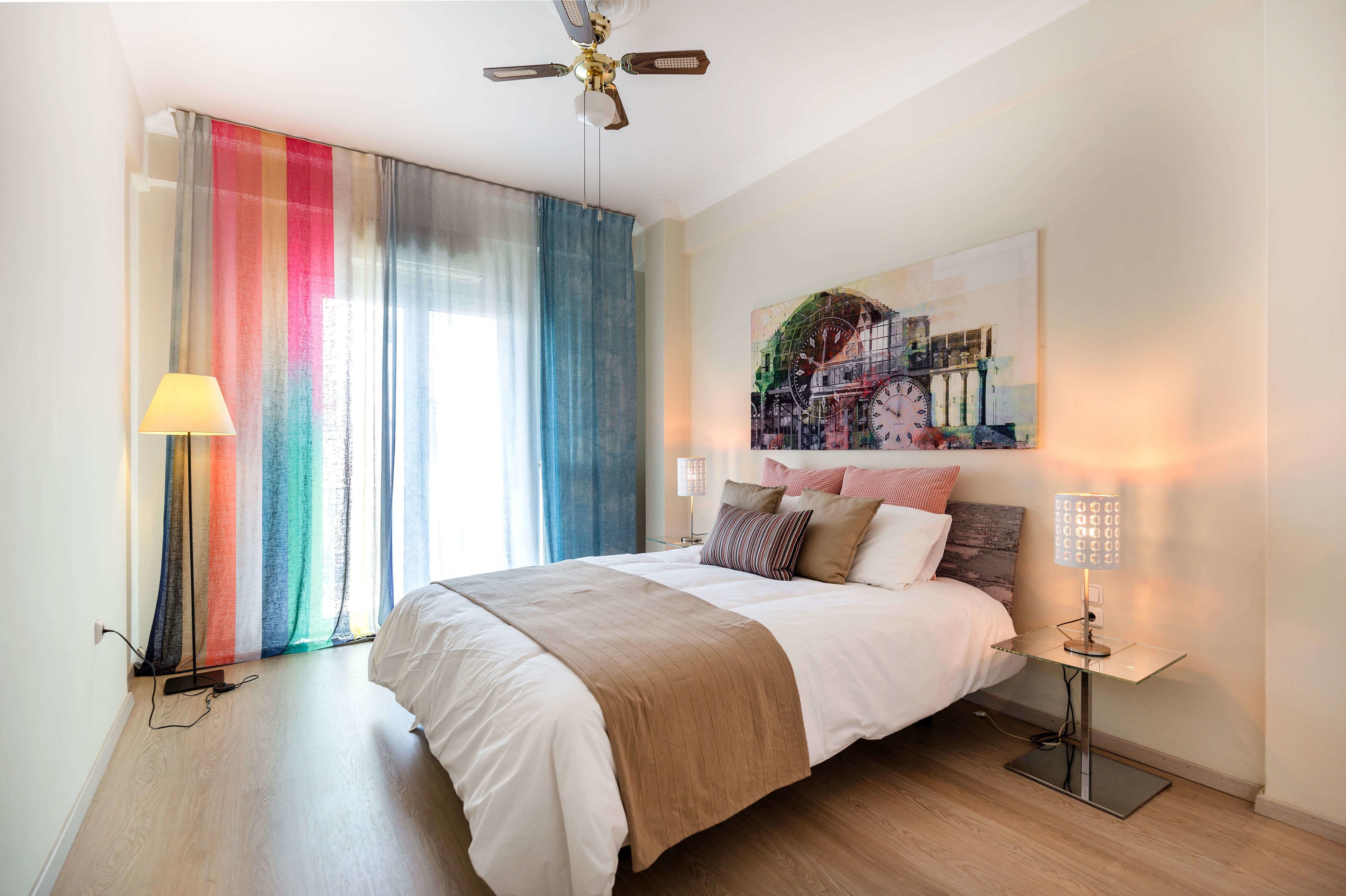 A bedroom with a bed, a ceiling fan, a lamp, and a painting on the wall.