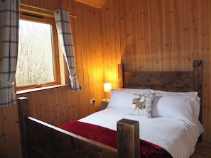 A wooden cabin bedroom with a bed, a window with curtains, a bedside lamp, and a red throw blanket. - The Cabins, Loch Awe (Taynuilt)