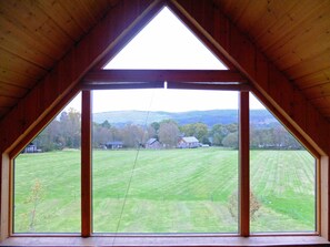 A room with a large triangular window offering a view of a green field and distant buildings. - The Cabins, Loch Awe (Taynuilt)