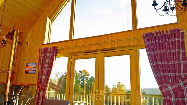 Una cabaña de madera con ventanas amplias, chimenea y cortinas de cuadros.