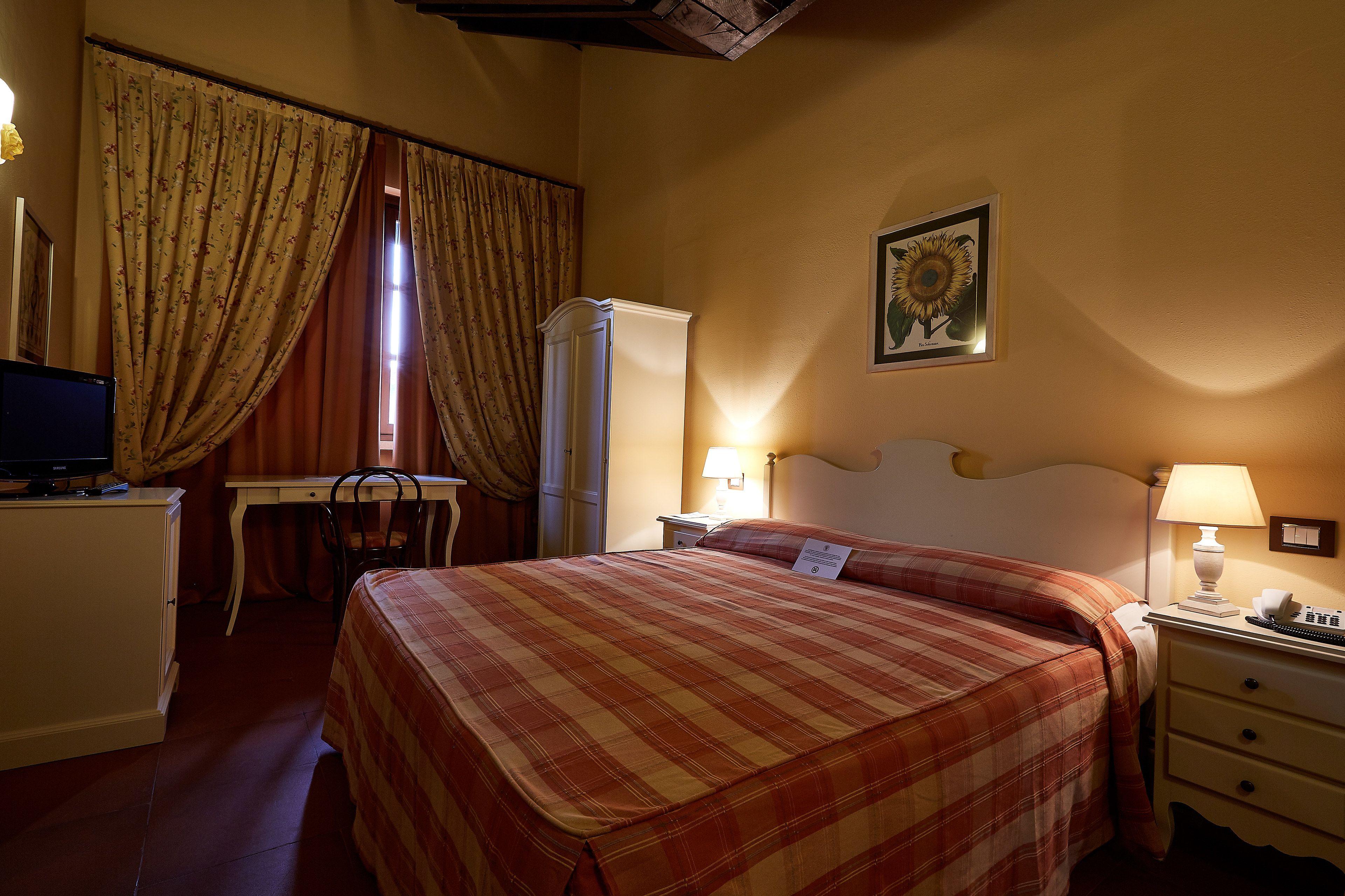 A hotel room with a bed, a desk, a chair, a wardrobe, and a nightstand with a lamp.