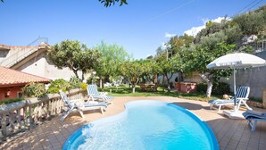 Una piscina con sillones de descanso y una sombrilla, rodeada de follaje y un muro de piedra.