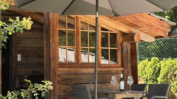 A wooden cabin with a dining area featuring a table and chairs, an umbrella, and a bottle of wine.