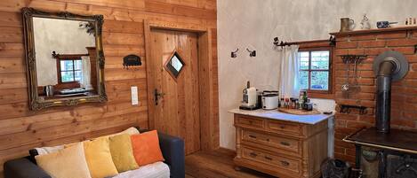 A cozy cabin interior with a wooden wall, a mirror, a sofa with colorful cushions, a wooden dresser, a kitchen area with a stove, and a window with a view of greenery.