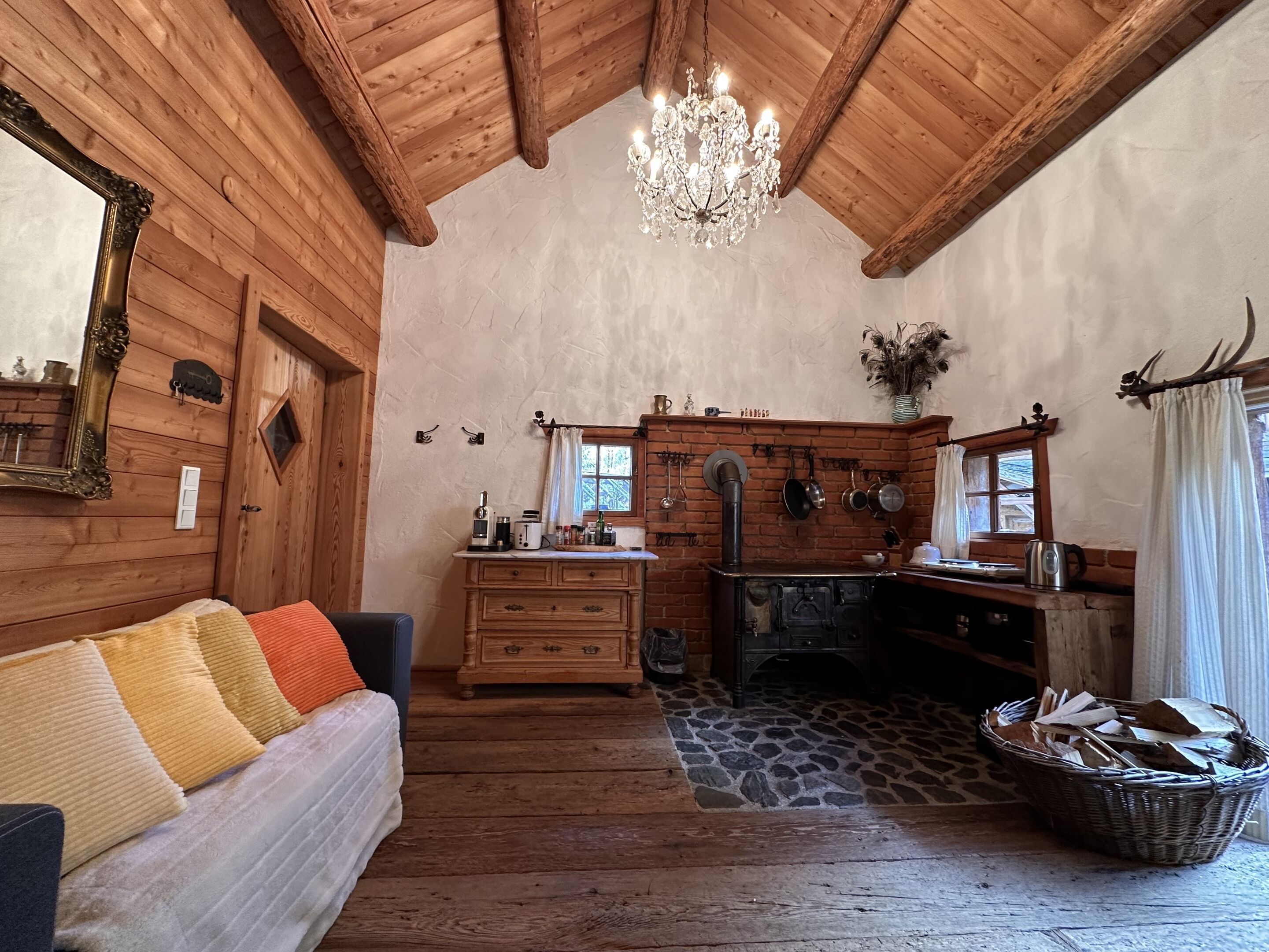 Interior acollidor d'una cabana de fusta, amb un sofà de fusta, un penjador de cristall, una llar de pedra i una zona de cuina amb olles penjades.