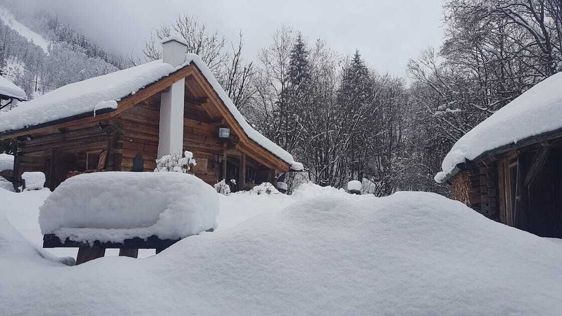 Una cabaña de madera con techo nevado y chimenea, rodeada por un paisaje invernal y árboles.