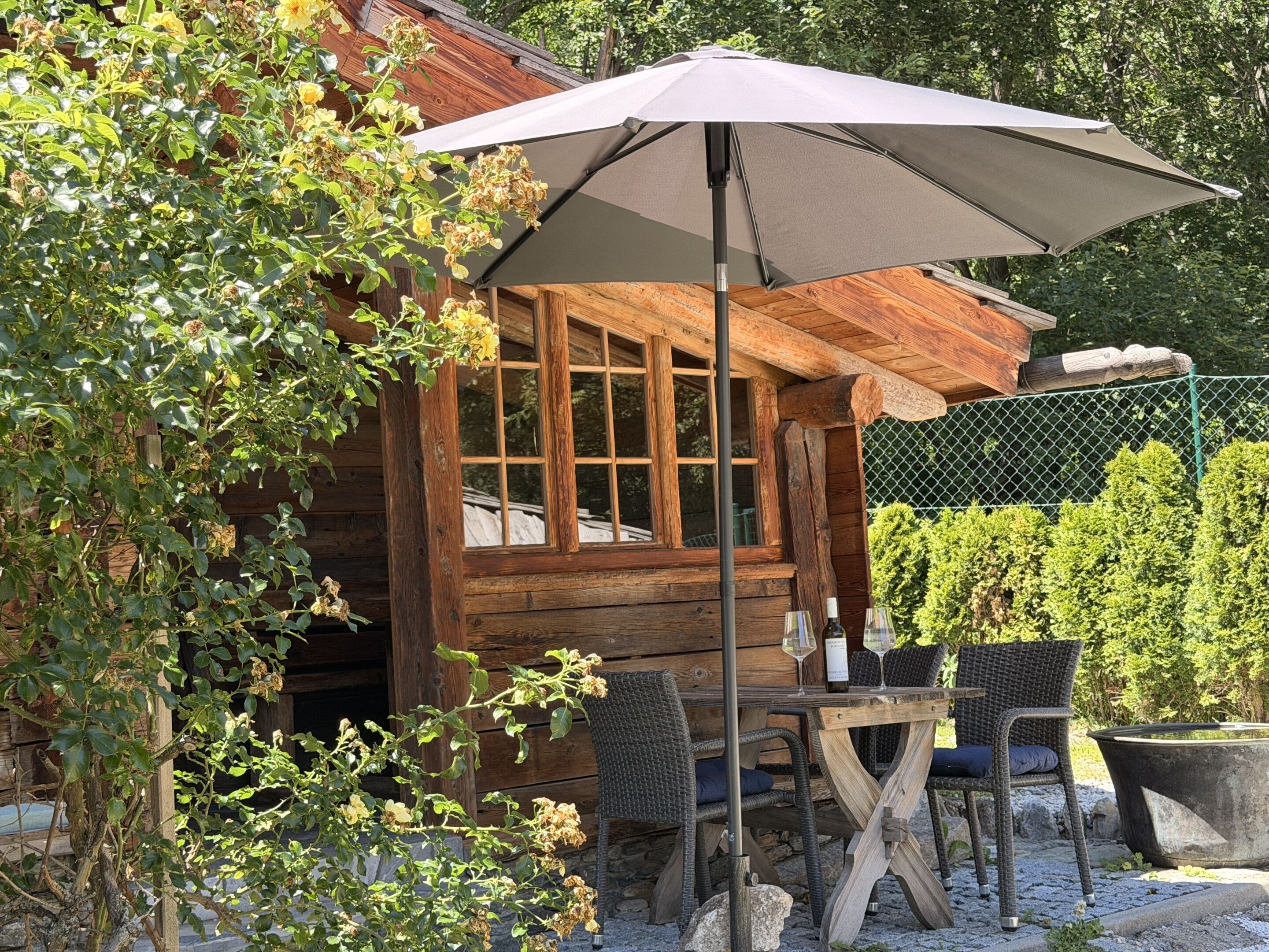 A wooden cabin with a table and chairs under a large umbrella, surrounded by greenery.
