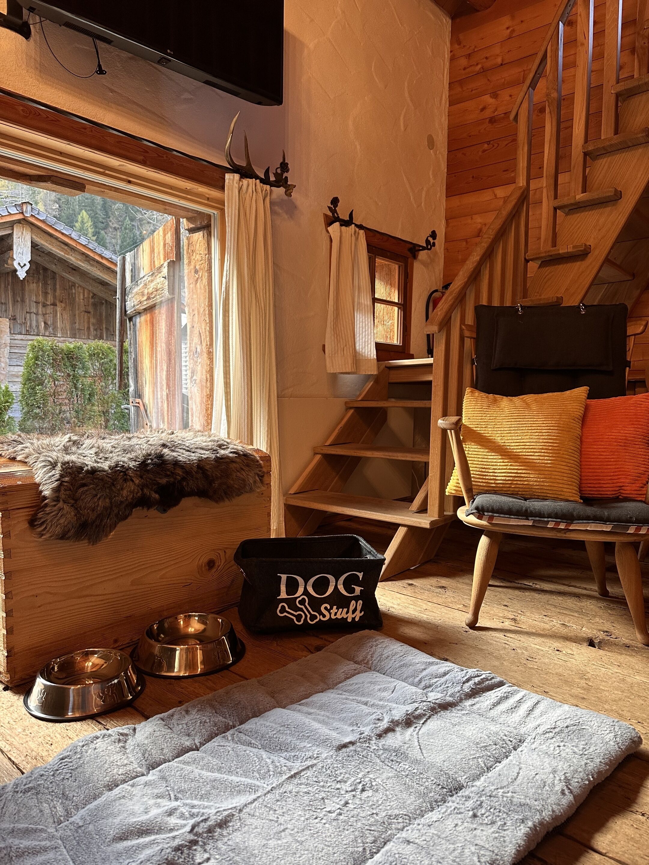 Interior acogedor de una cabaña de madera con una cama para perro, una escalera y una ventana con vistas a un bosque.