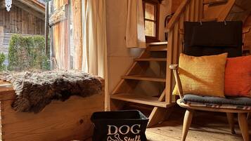 Interior acogedor de una cabaña de madera con una cama para perro, una escalera y una ventana con vistas a un bosque.