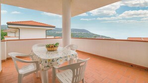 Un balcone con tavolo e sedie, un cesto di vimini pieno di frutta e vista su una città e montagne.