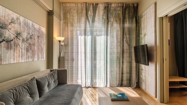 Una sala de estar moderna con un sofá, una mesa de centro, un televisor y ventanas amplias con cortinas translúcidas.